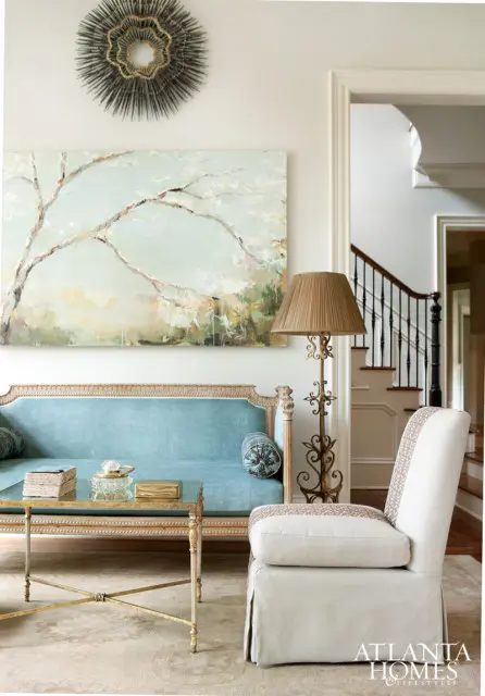 Living room with a light blue velvet sofa, ornate gold coffee table, and a slipcovered armchair. A large abstract painting hangs above the sofa, and a metal sunburst mirror is on the wall.