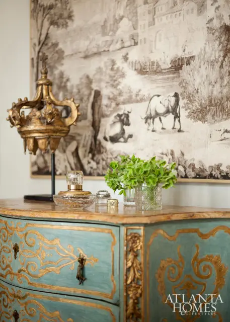 Turquoise bombe chest with ornate gold scrollwork and brass hardware, topped with a gold crown finial, crystal perfume bottle, and a glass vase of greenery.