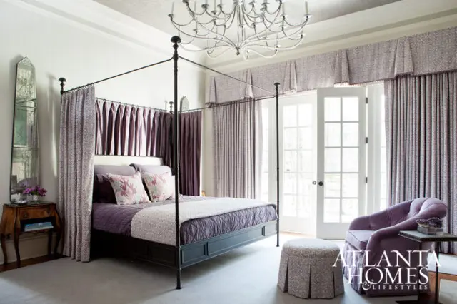 Bedroom with a four-poster bed draped in purple fabric, matching curtains, a tufted purple armchair, and a crystal chandelier.