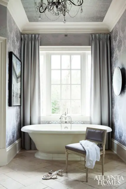 Cream freestanding bathtub with chrome fixtures in a bathroom with grey damask wallpaper, grey drapes, and a crystal chandelier.