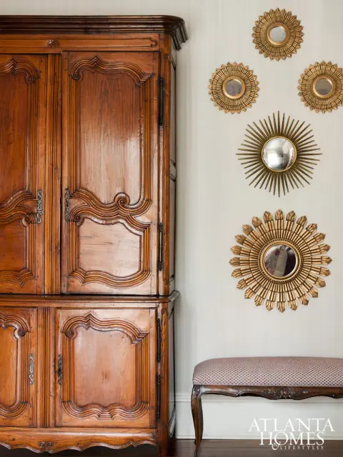Antique wooden armoire with carved details, a bench with patterned upholstery, and a collection of sunburst mirrors on a textured wall.