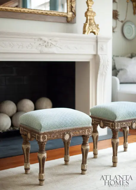 Two light blue upholstered ottomans with carved wooden legs in front of a white fireplace with a gold mirror above
