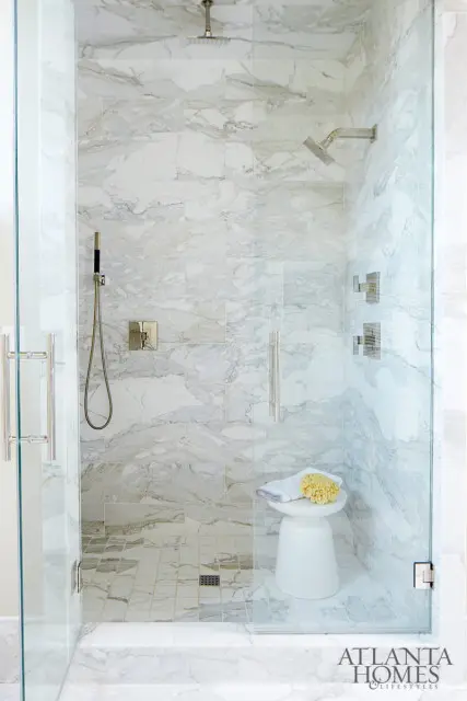 Glass-enclosed shower with white marble tile walls and floor, brushed nickel fixtures, and a small white stool with a towel and loofah.