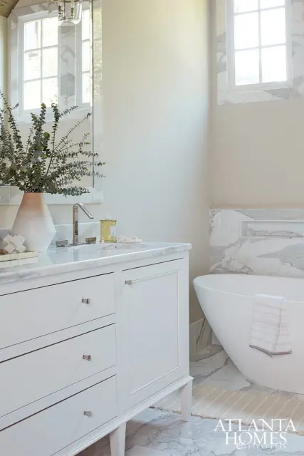 White bathroom vanity with marble countertop, chrome faucet, and a vase of eucalyptus. Freestanding tub with marble tile surround and a striped towel visible.