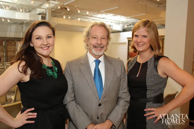 Three people smiling at the camera: a woman in a black dress with a green necklace, a man in a grey suit and blue tie, and a woman in a black and grey dress.