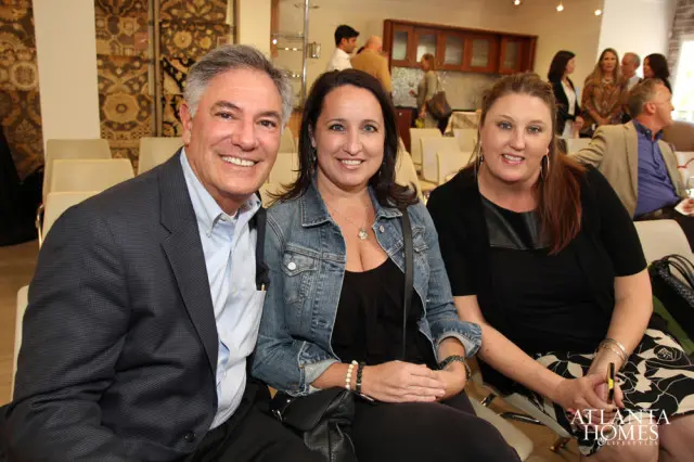 Man in a dark suit and light blue shirt smiles next to two women, one in a denim jacket and the other in a black cardigan.