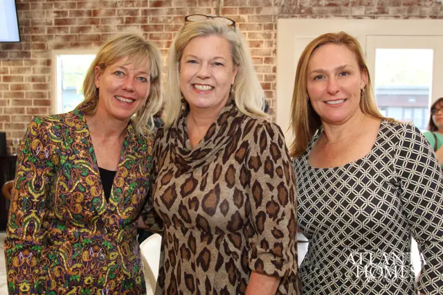 Three women smiling at the camera. The woman on the left wears a colorful paisley jacket, the woman in the middle wears a leopard print dress, and the woman on the right wears a black and white geometric print dress.
