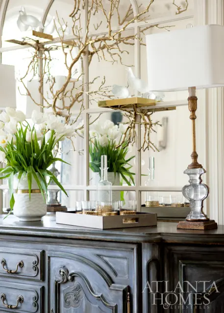 Dark distressed buffet topped with a white vase of tulips, a tray of glasses and a decanter, a lamp, and a gold branch with glass birds against a mirrored wall.