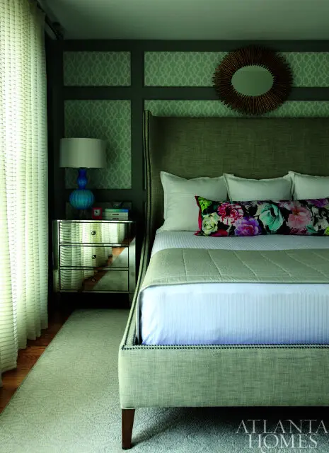 Bedroom with a tall upholstered headboard, white bedding, and a floral bolster pillow. A mirrored nightstand holds a blue lamp, and a sunburst mirror hangs on the wall.