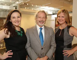 Two women and a man stand together, smiling. The woman on the left wears a black dress and a green necklace. The man in the center wears a gray suit and blue tie. The woman on the right wears a black and gray patterned dress.
