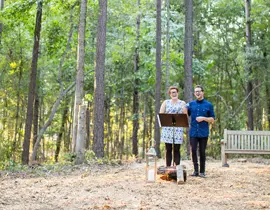 Two people stand in a clearing in a wooded area with a wooden bench in the background. One person stands behind a music stand with a microphone.