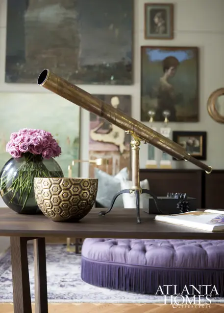 Dark wood table with brass telescope, glass vase of pink roses, gold honeycomb bowl, and tufted purple ottoman with fringe.