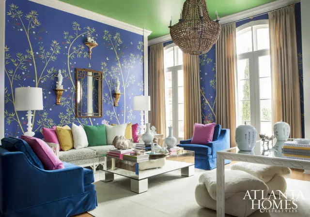 Living room with blue Chinoiserie wallpaper, green ceiling, and plush blue velvet seating. A mirrored coffee table holds decorative objects and books.