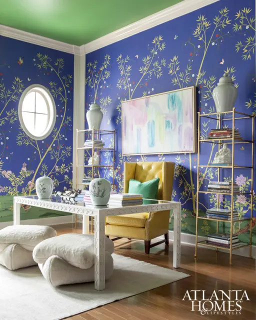 Blue Chinoiserie wallpaper adorns the walls of a room with a green ceiling, featuring a yellow tufted armchair, a white coffee table with decorative vases, and gold shelving units.