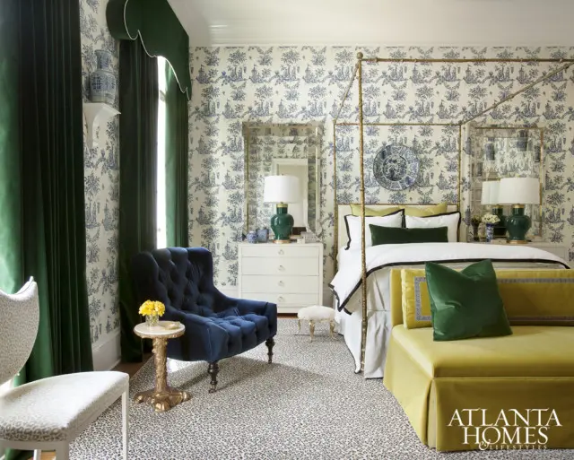 Bedroom with blue and white toile wallpaper, a brass four-poster bed, green velvet curtains, a navy blue tufted armchair, and a yellow velvet chaise lounge.
