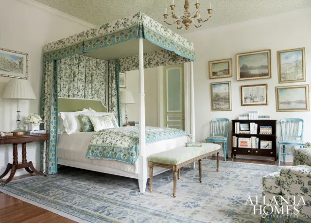 Bedroom with a four-poster bed draped in floral fabric, a green upholstered headboard, and matching bedding. A patterned rug, light blue chairs, and artwork adorn the space.
