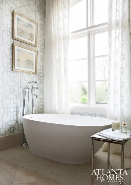 Freestanding white bathtub with chrome faucet against a wall with light blue geometric wallpaper and two framed landscape paintings. Sheer white curtains frame a large window.