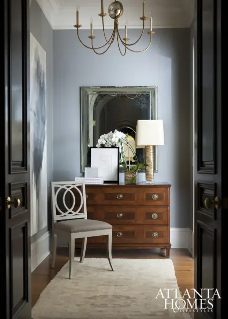 Hallway with gray walls, a wooden dresser, a chair with a geometric back, and a gold chandelier.