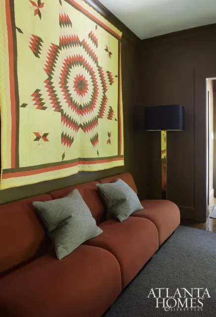 Living room with a rust-colored sofa, two gray patterned pillows, a large starburst quilt hanging on a dark brown wall, and a tall black-shaded floor lamp.