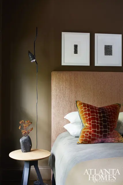 Bedroom with a textured coral headboard, gray bedding, and an orange and red patterned accent pillow. A black floor lamp stands beside a small wooden side table with a dark vase holding dried flowers.