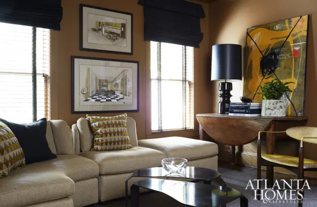 Living room with a sectional sofa, geometric patterned pillows, framed architectural drawings, a drop-leaf table with a black lamp and abstract art, and a logo for Atlanta Homes & Lifestyles.
