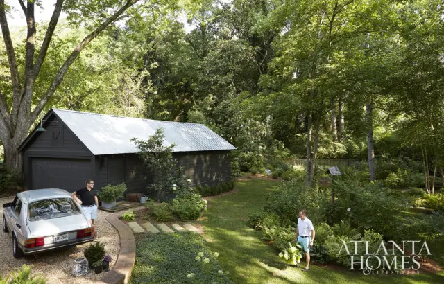Dark wood garage with a silver metal roof, parked next to a silver vintage car. A man stands by the car, and another man walks through a lush, wooded backyard with a birdhouse.
