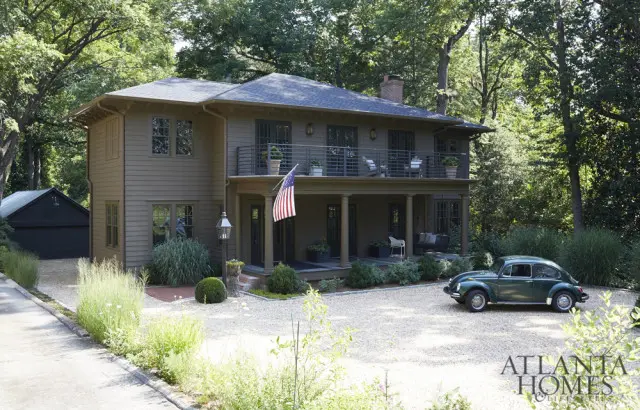 Two-story tan house with a wraparound porch, balconies, and a dark garage in the background. A vintage green Volkswagen Beetle is parked in the gravel driveway.