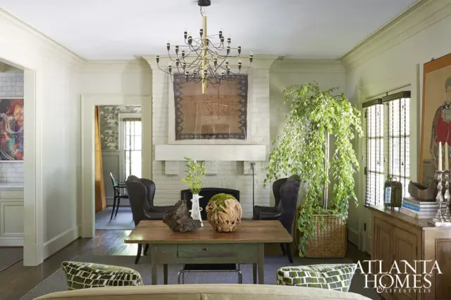 Living room with a white brick fireplace, a large green tree, dark leather chairs, and a rustic wooden table with decorative objects.