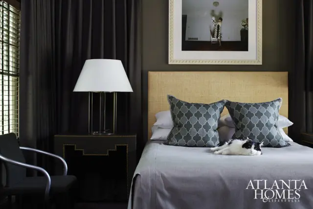 Bedroom with a woven headboard, grey bedding, and decorative pillows. A black and white cat rests on the bed. Dark curtains frame a window with blinds.