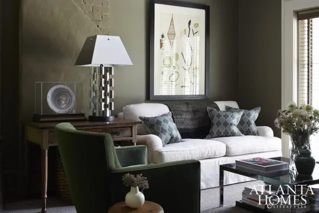 Living room with a white slipcovered sofa, green armchair, dark wood side table with a metallic lamp, and framed abstract art on a dark green wall.
