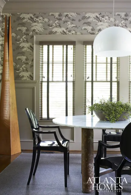 Dining room with patterned wallpaper, marble-top table with a plant, black chairs, and a tall wooden sculpture.