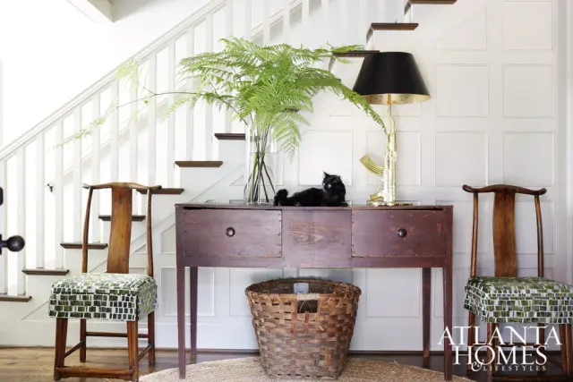Entryway with a dark wood console table, two wooden chairs with green mosaic patterned seats, a black cat, a large fern, and a gold lamp shaped like a rifle.