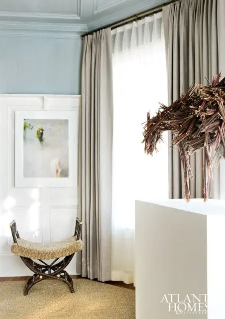 Light blue paneled ceiling and walls with a framed abstract painting and a twig sculpture. A woven bench sits on a jute rug in front of floor-to-ceiling curtains.