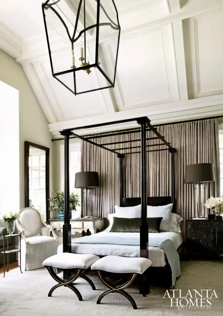 Bedroom with a black canopy bed, white bedding, and olive green pillows. Flanked by bedside tables with dark lamps and a large window with pleated drapes behind. A large black lantern hangs from the coffered ceiling.