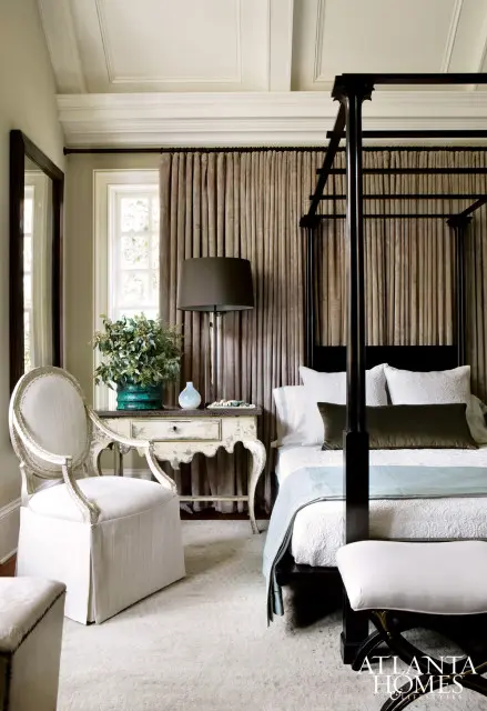 Bedroom with a dark wood four-poster bed, white bedding, and a light blue throw. A white upholstered chair sits next to a distressed antique table with a lamp and plant.