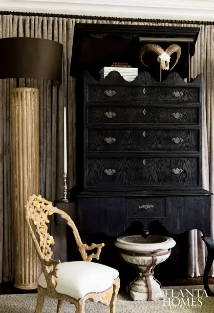 Dark wood dresser with ornate hardware, a ram skull, and books, next to a carved chair with a white cushion and a fluted column.