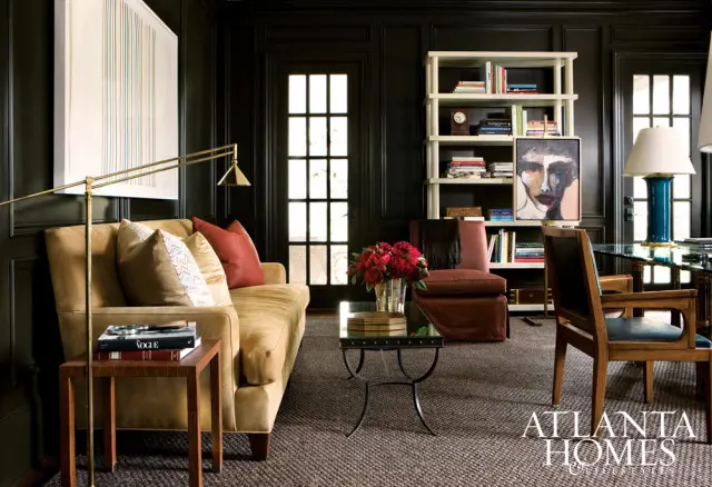 Dark paneled living room with a tan sofa, red and patterned pillows, a glass-top coffee table with a floral arrangement, a bookshelf filled with books and art, and a wooden chair at a desk.