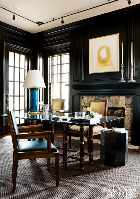 Dark paneled office with glass-top desk, blue lamp, stone fireplace, and artwork above the mantel.