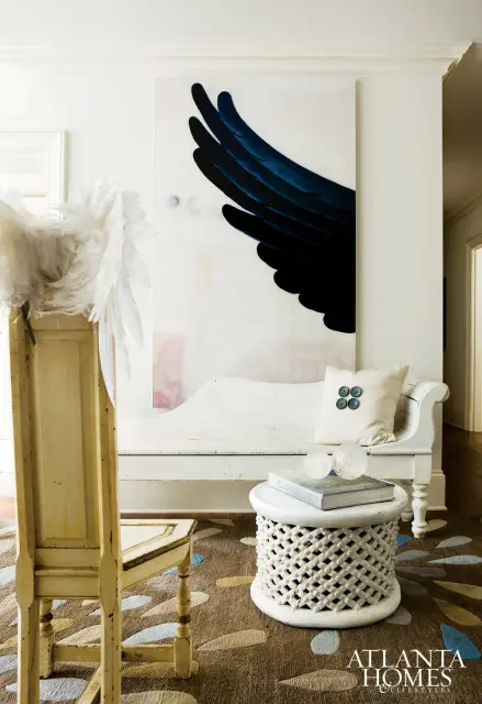 White bench with a cream pillow featuring four dark buttons, next to a white carved side table with books and glass spheres. A large painting of a dark blue feather wing hangs on the wall.