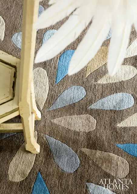 Close-up of a textured rug with a pattern of teardrop shapes in beige, light blue, and gray on a brown background. A portion of a cream-colored wooden chair leg is visible on the left.