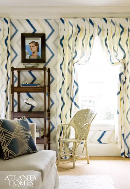 Living room with a white sofa, wicker chair, and patterned curtains and wallpaper in blue, yellow, and gray. A wooden bookshelf holds decorative items.
