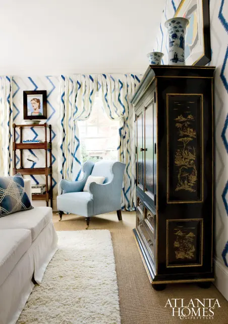 Living room with blue and white ikat patterned wallpaper, matching curtains, a light blue wingback chair, a white sofa, a dark lacquered cabinet with gold chinoiserie details, and a shag rug.