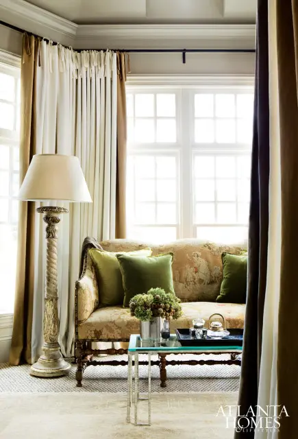 Living room with a tapestry-upholstered sofa, green velvet pillows, a vintage floor lamp, and a glass-top coffee table with a floral arrangement.