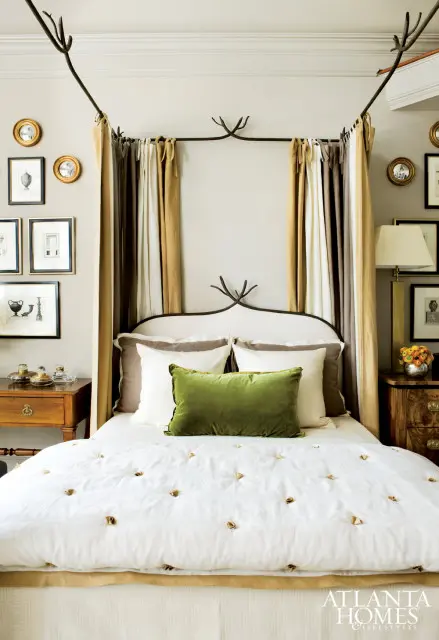 Bedroom with a white upholstered headboard and a dark metal canopy frame. Drapes in beige and brown hang from the canopy. A white duvet with gold accents covers the bed, topped with a green velvet lumbar pillow.