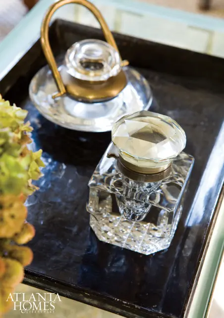 Close-up of antique crystal inkwell with faceted lid and brass accents on a dark tray, next to a decorative glass object with a brass handle and a sprig of dried flowers.