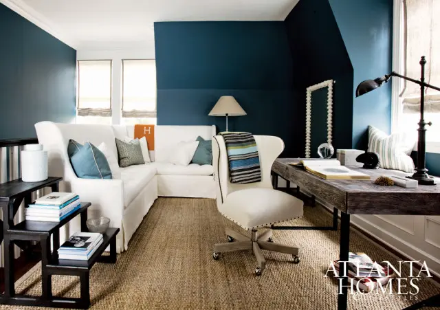 Living room with deep blue walls, a white sectional sofa, a white upholstered desk chair, and a dark wood desk. A woven rug covers the floor.