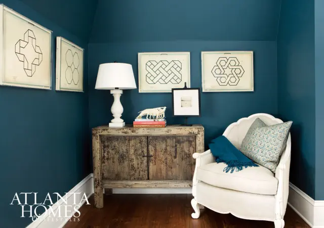 Corner of a room with deep teal walls, featuring a distressed wooden cabinet, a white armchair with patterned pillows and a teal throw, and framed geometric art.