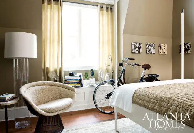 Bedroom with a white bed, quilted brown comforter, and a white throw. A textured beige armchair sits next to a glass and metal floor lamp. A bicycle is parked near a window with beige curtains.