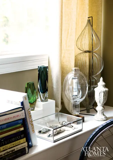 Stack of books including Louis Vuitton Impressions, and decorative objects including glass vases, a large Edison bulb lamp, and a wire sculpture on a windowsill with yellow curtains.