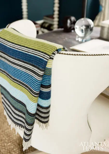 White upholstered chair with a striped throw blanket in shades of blue, green, and black draped over the back. A wooden desk with decorative objects is visible in the background.